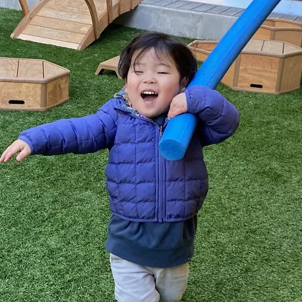 A smiling child in a blue puffer jacket holds a blue foam tube over one shoulder while standing on green artificial turf near wooden play structures.