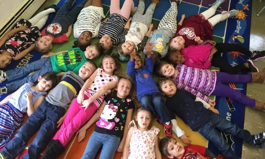 A group of children lie on their backs in a circle on a colorful alphabet rug, looking up at the camera.