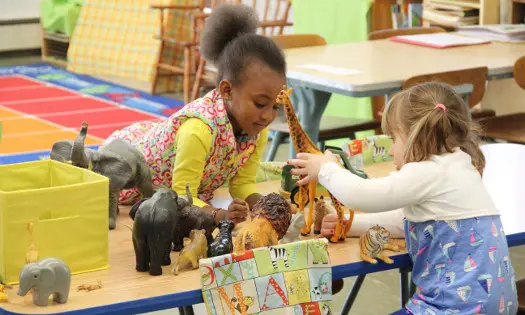 Two young girls stand at a classroom table playing with toy animals including elephants, a giraffe, and a lion.
