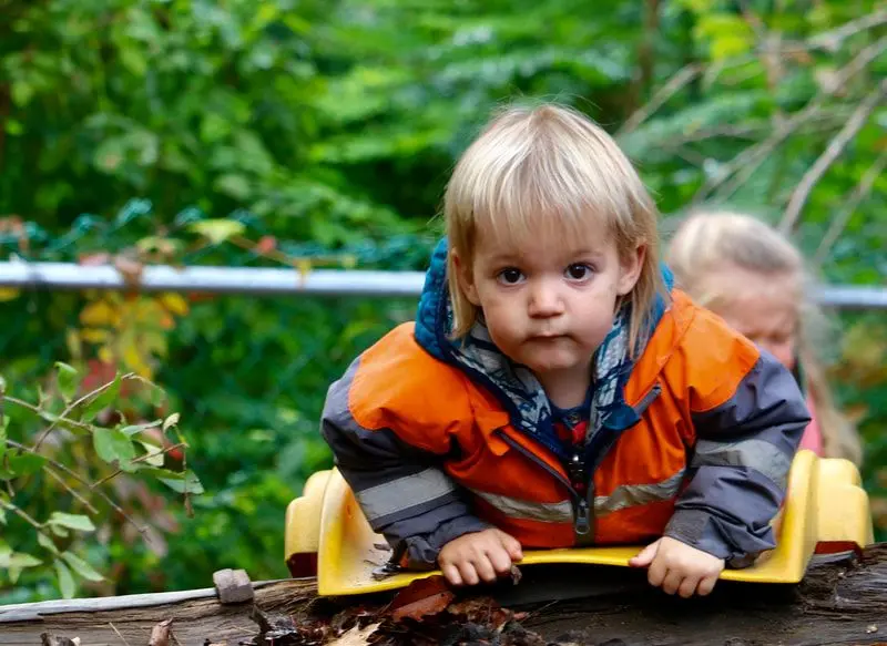A young child with blonde hair in an orange and grey jacket leans forward on a yellow toy while another child sits behind among green foliage.