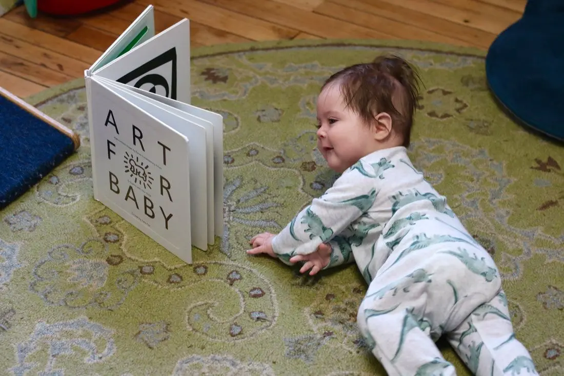 A baby in patterned pajamas lies on a rug and looks at an open board book titled ART FOR BABY.