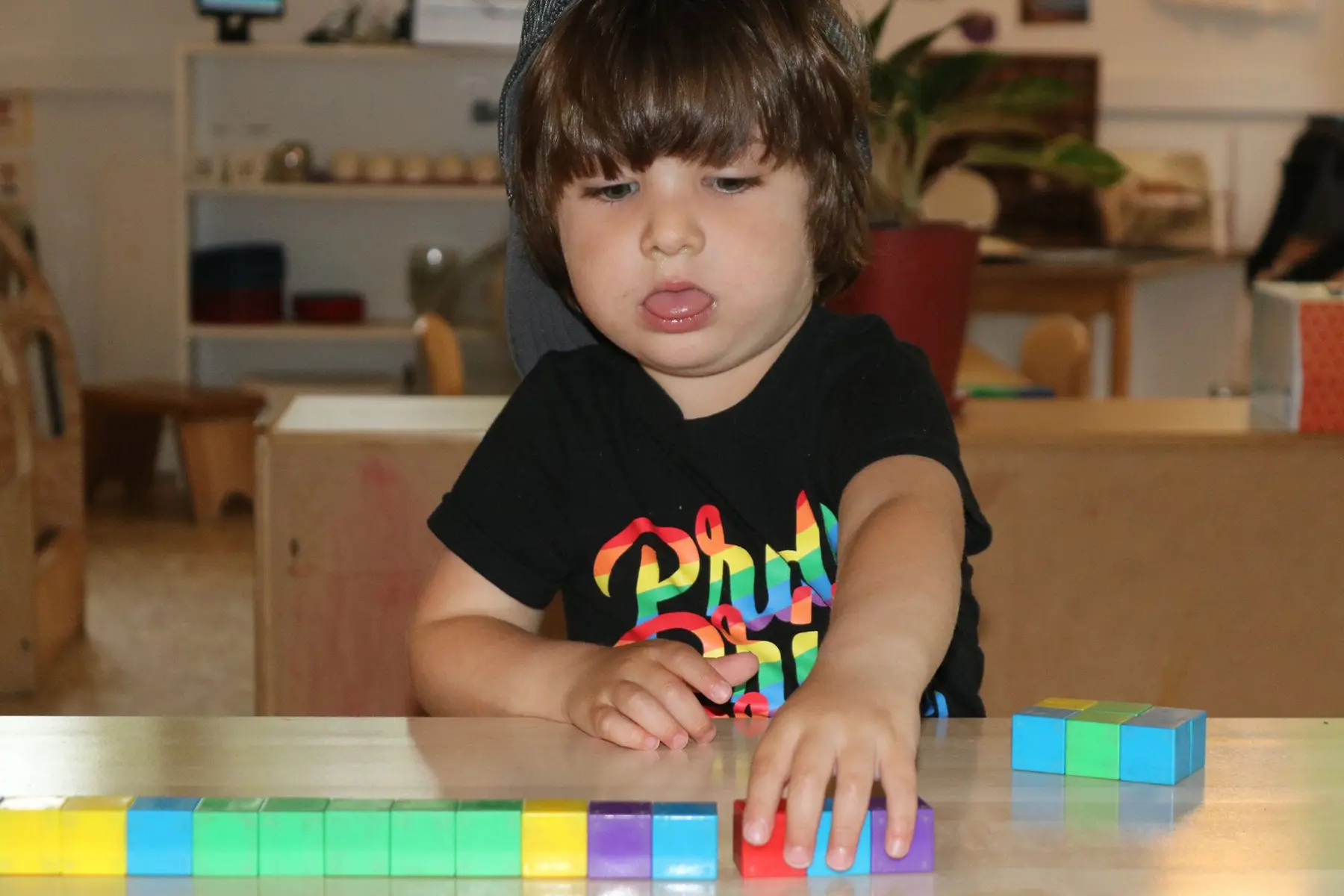 A young child in a black graphic t-shirt stands at a table, touching a row of colorful connected cubes with one hand while looking down.