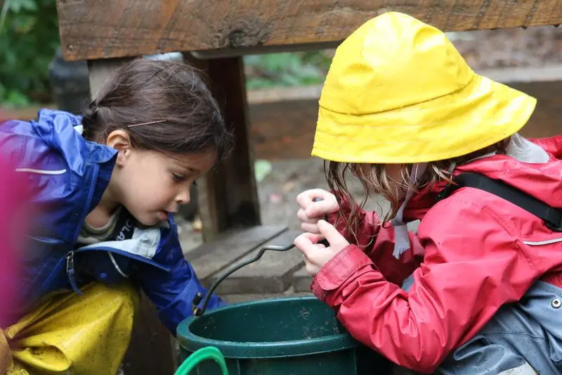Two children in rain jackets and a yellow hat play with a metal pump and a green bucket outdoors.