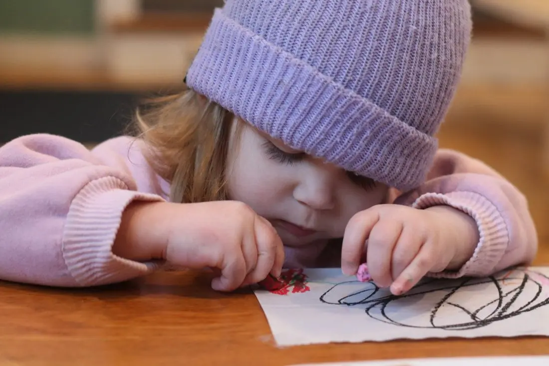 A young child wearing a purple knit beanie and pink sweater rests their head on a table while drawing with a crayon on paper.