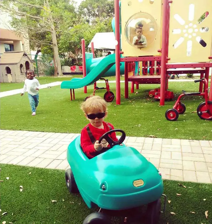 A child in a red shirt and sunglasses sits in a teal toy car on artificial grass while another child walks on the grass behind near a play structure.