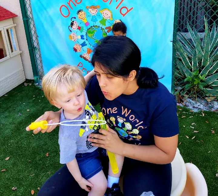 A woman in a dark blue 'One World' shirt sits on a yellow chair and blows bubbles with a wand while a young child with blonde hair reaches toward the bubbles.