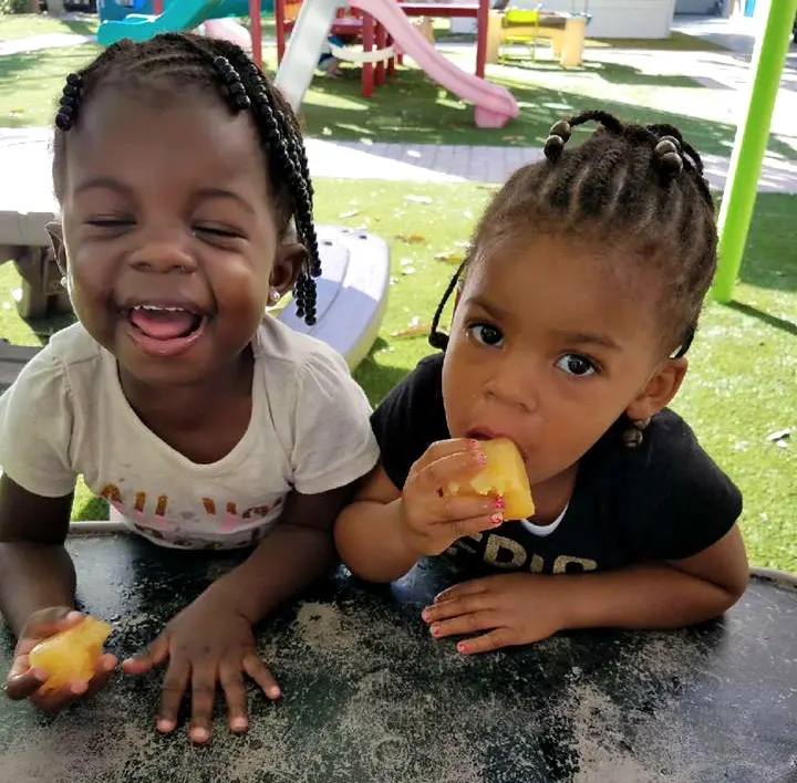 Two young girls sit at a dark table outdoors, one laughing with an open mouth and the other eating a piece of yellow fruit.