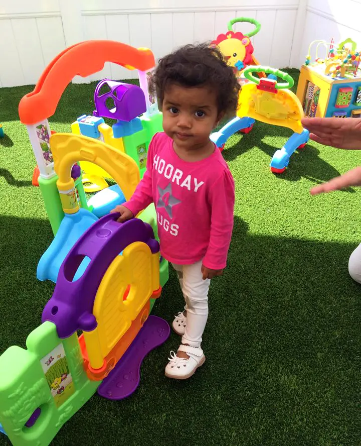 A toddler in a pink shirt stands beside a colorful plastic activity center on artificial grass while an adult hand reaches toward a toy in the background.