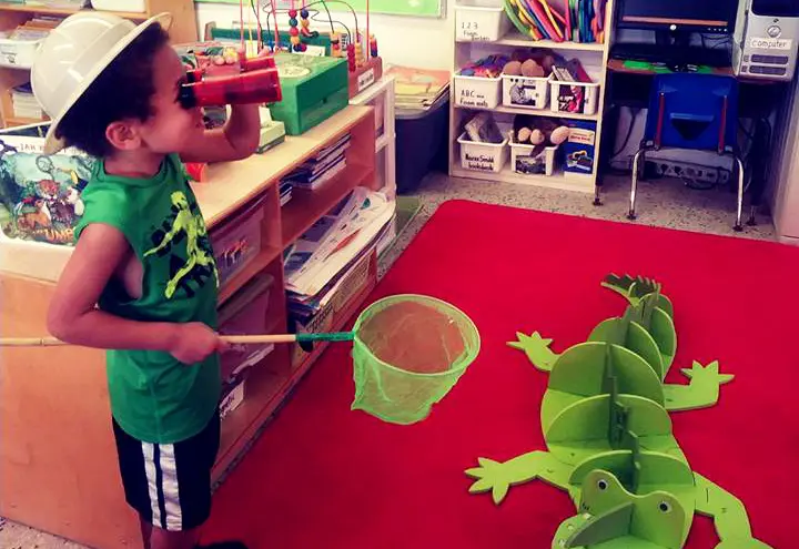 A young boy in a green shirt and white hat drinks from a red cup while holding a green net near a large green foam alligator on a red mat in a classroom.