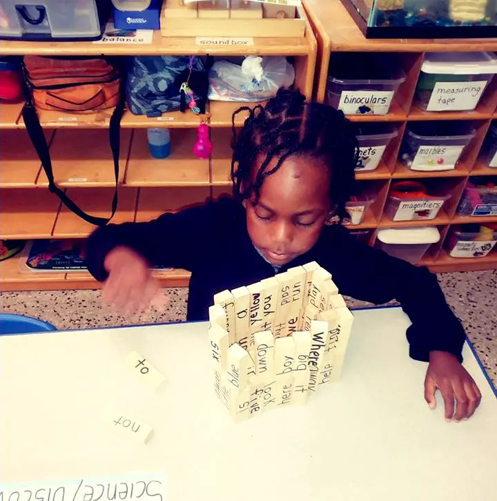 A child with braided hair sits at a white table and builds a tower from wooden blocks with printed words while looking down at the blocks.