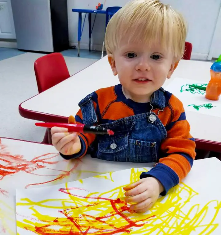 A young child in a striped shirt and denim overalls holds a red marker while sitting at a table with paper covered in red and yellow scribbles.