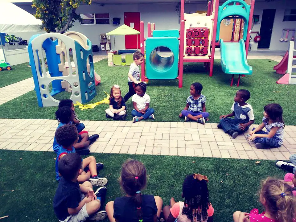 Children sit on a paved and artificial-grass play area while one child walks near a blue and teal play structure with a slide.