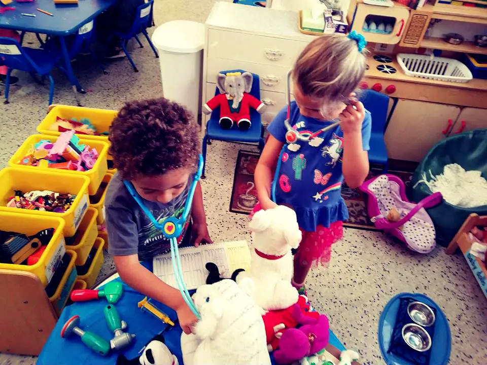 Two children play with stuffed animals and toy medical tools at a blue table in a classroom with yellow bins and play furniture.