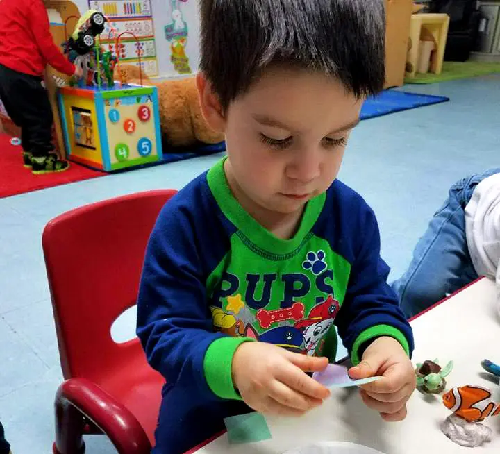 A young boy in a blue and green PAW Patrol shirt sits at a table holding a small piece of paper while another child in a red shirt stands near a toy cube in the background.