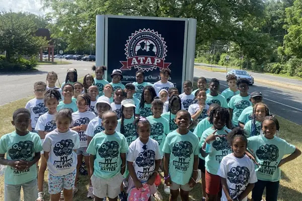 A large group of children wearing matching light green t-shirts stands together on a paved area beside a road and a large dark sign.