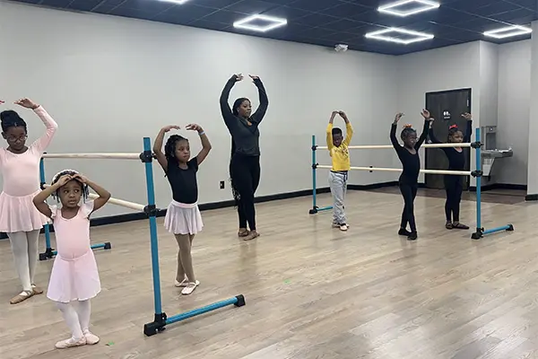 Five children and one adult stand in a dance studio with blue barres, most holding their arms above their heads.