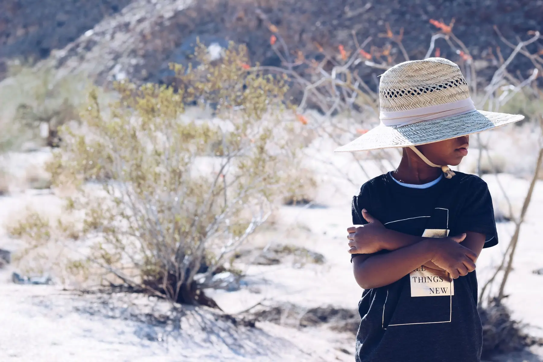 A young child wearing a wide-brimmed straw hat and a black t-shirt stands with arms crossed in a dry, sunlit desert landscape with sparse bushes.