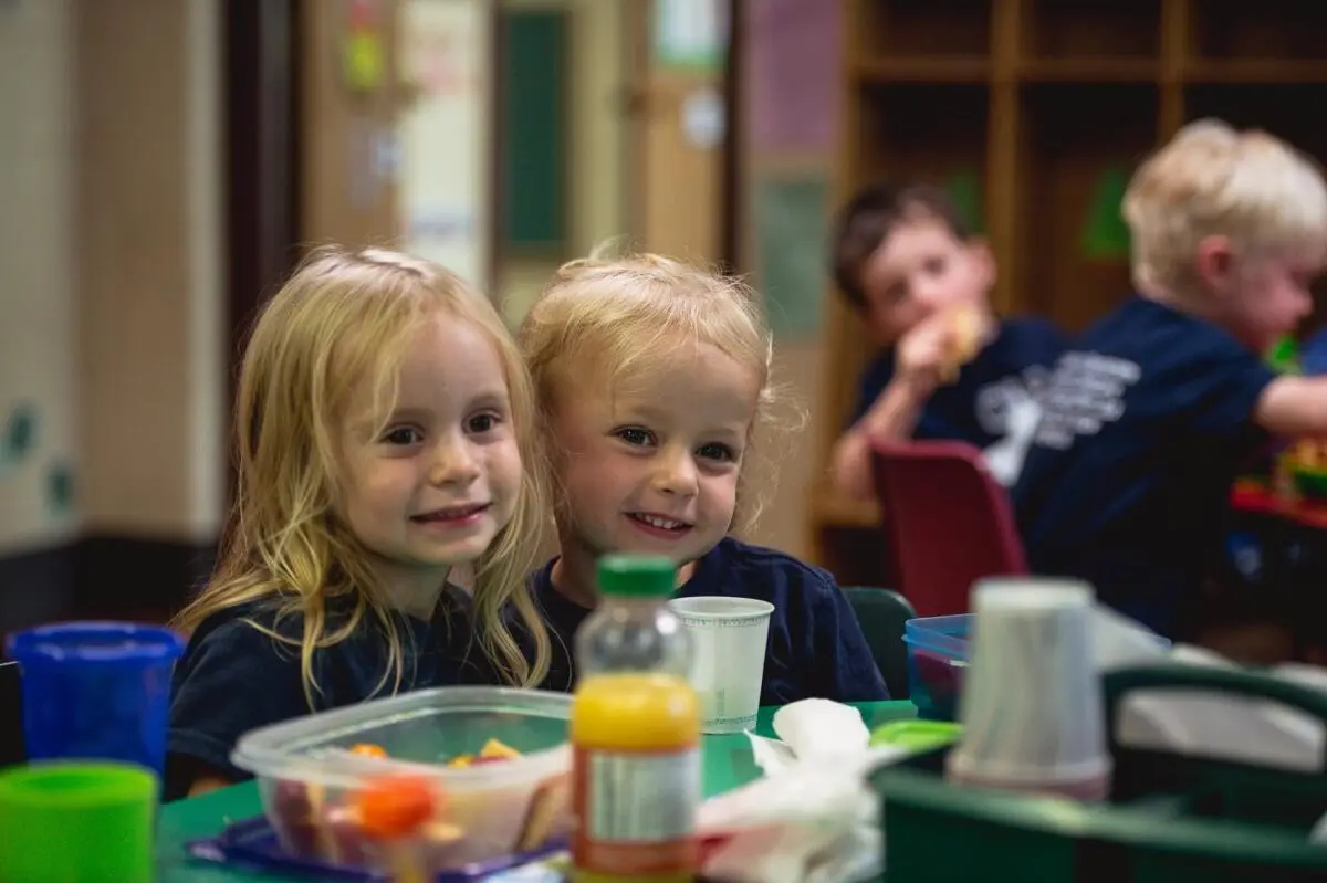 Two young girls with blonde hair sit at a table with food containers and cups while two boys eat in the background.