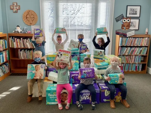 Eight children wearing face masks sit and stand among stacks of diaper packages in a room with bookshelves and a window.