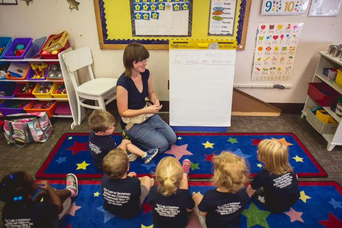 A woman sits on a star-patterned rug facing six children seated in a semicircle beside a whiteboard and alphabet poster in a classroom.