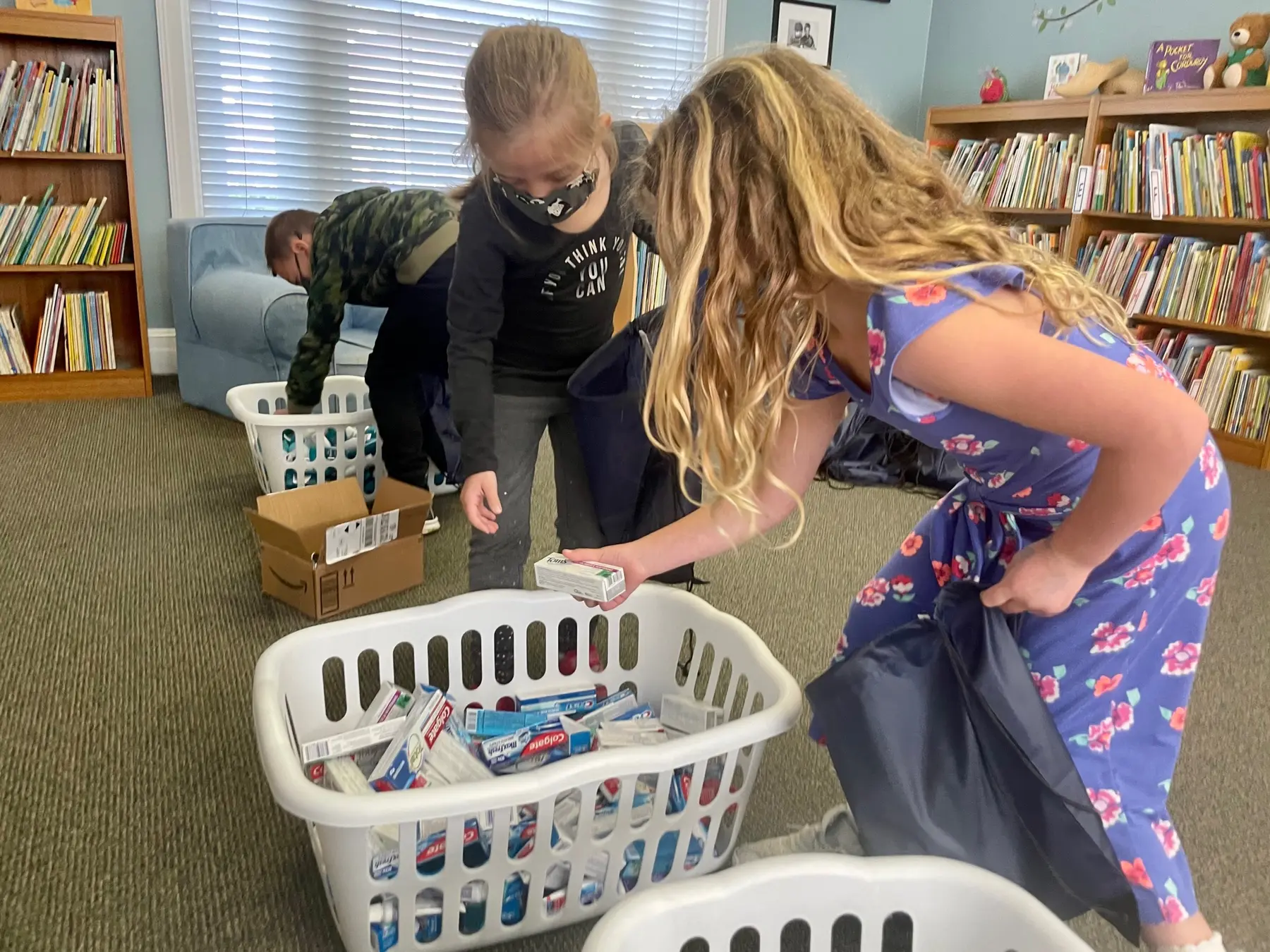 Three children stand around white laundry baskets filled with boxed items in a room with bookshelves, as one child in a floral dress places a box into a basket.