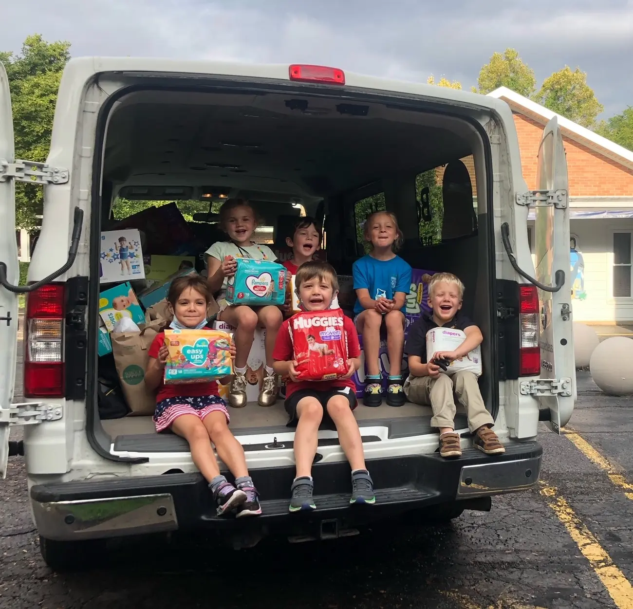 Five children sit in the open back of a white van, holding packages of diapers and wipes while smiling.