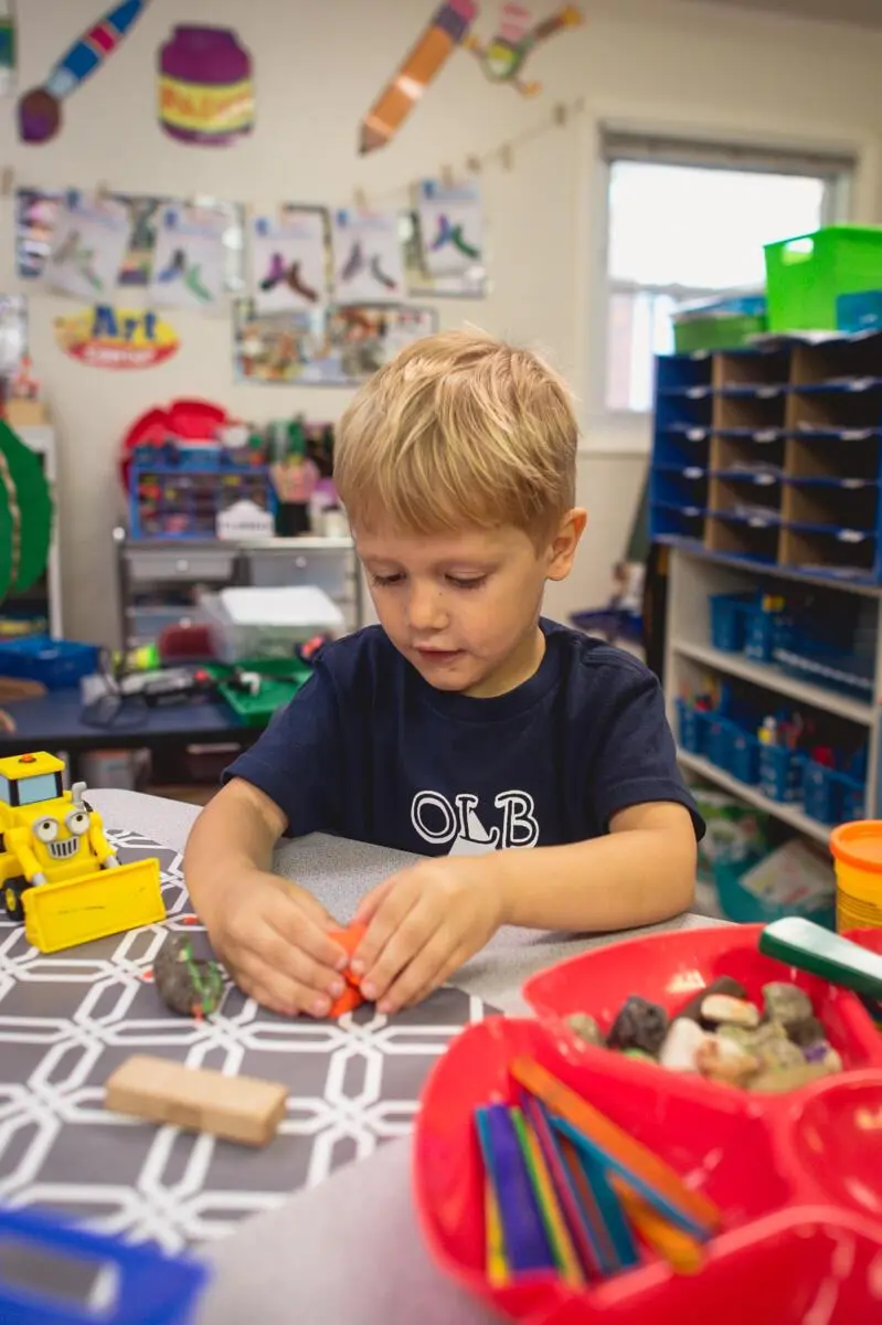 A young boy in a dark blue shirt sits at a table and manipulates a small orange object beside a yellow toy truck and red bins of art supplies.