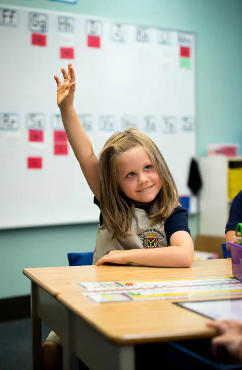 A girl with shoulder-length blonde hair sits at a wooden desk with her arm raised, smiling, in front of a whiteboard with papers.