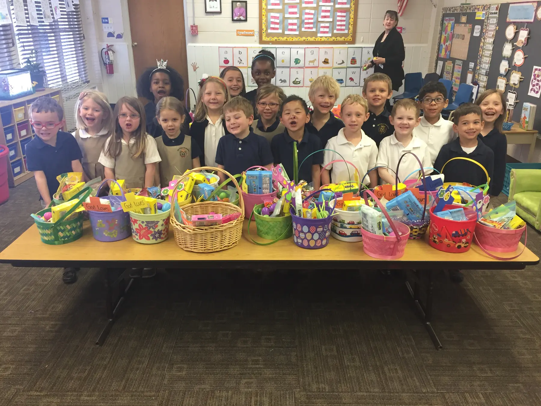 A group of children stand behind a long table displaying decorated Easter baskets filled with items, while an adult stands near the back wall of a classroom.