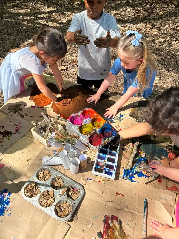 Four children stand and reach around a table covered with paper, paint cups, brushes, and trays of brown materials while painting outdoors.