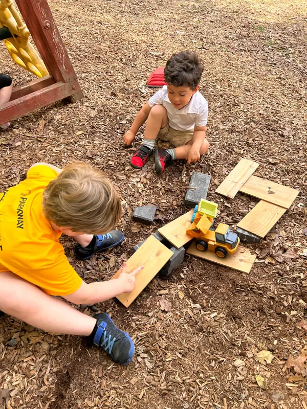 Two boys sit on wood chips playing with a toy construction truck and wooden planks.