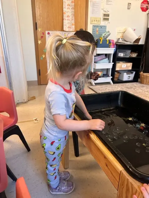 A young girl with a ponytail stands at a black water table and touches the water while another child sits behind it in a classroom.