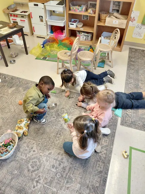 Five children sit or lie on a patterned rug in a classroom, playing with small toys and containers near shelves and a play kitchen.