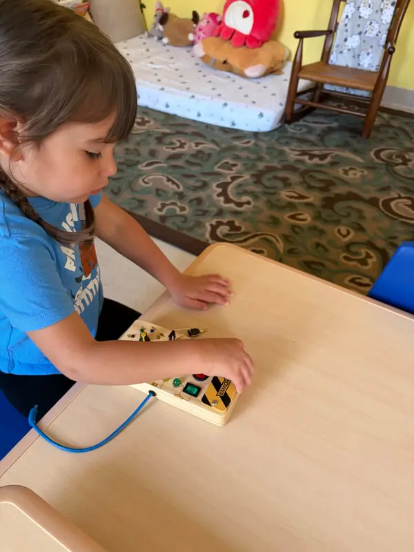 A young girl in a blue shirt stands at a light wood table and presses a button on a yellow electronic toy with a blue cord.