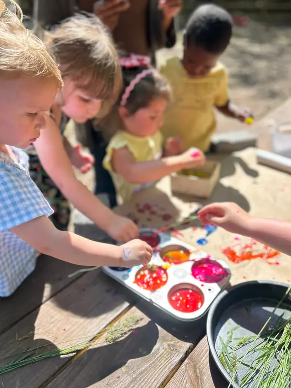 Several children are gathered around a wooden table outdoors, engaged in a craft activity with colorful liquids in a muffin tin and natural elements.