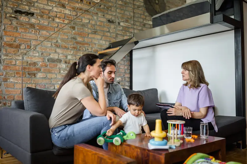 Two adults sit on a dark sofa talking with a woman in a purple top while a toddler plays with a green toy car on a wooden coffee table holding stacking toys and glasses.