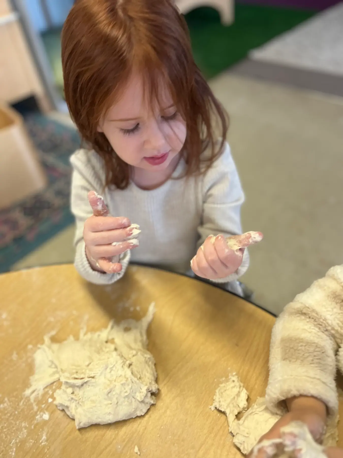 A child with red hair sits at a table and holds pieces of dough while another child in a fuzzy sleeve plays with dough nearby.
