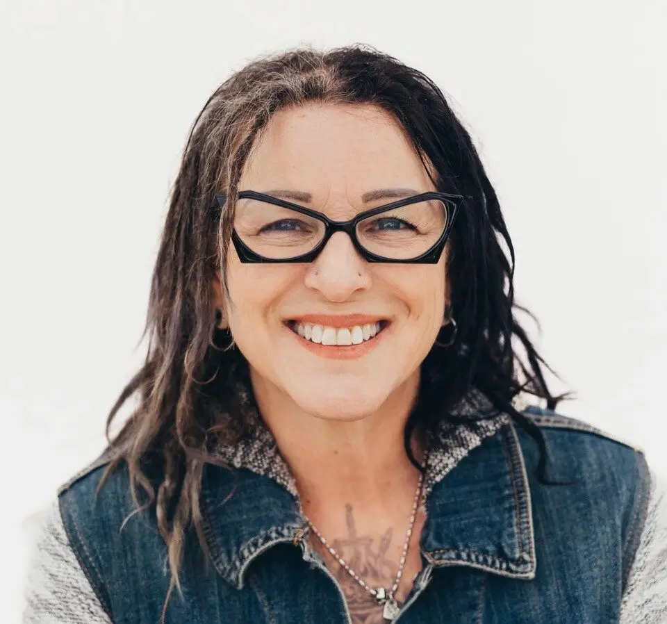 A smiling woman with long dark hair, black cat-eye glasses, a nose stud, and a denim vest stands against a plain white background.