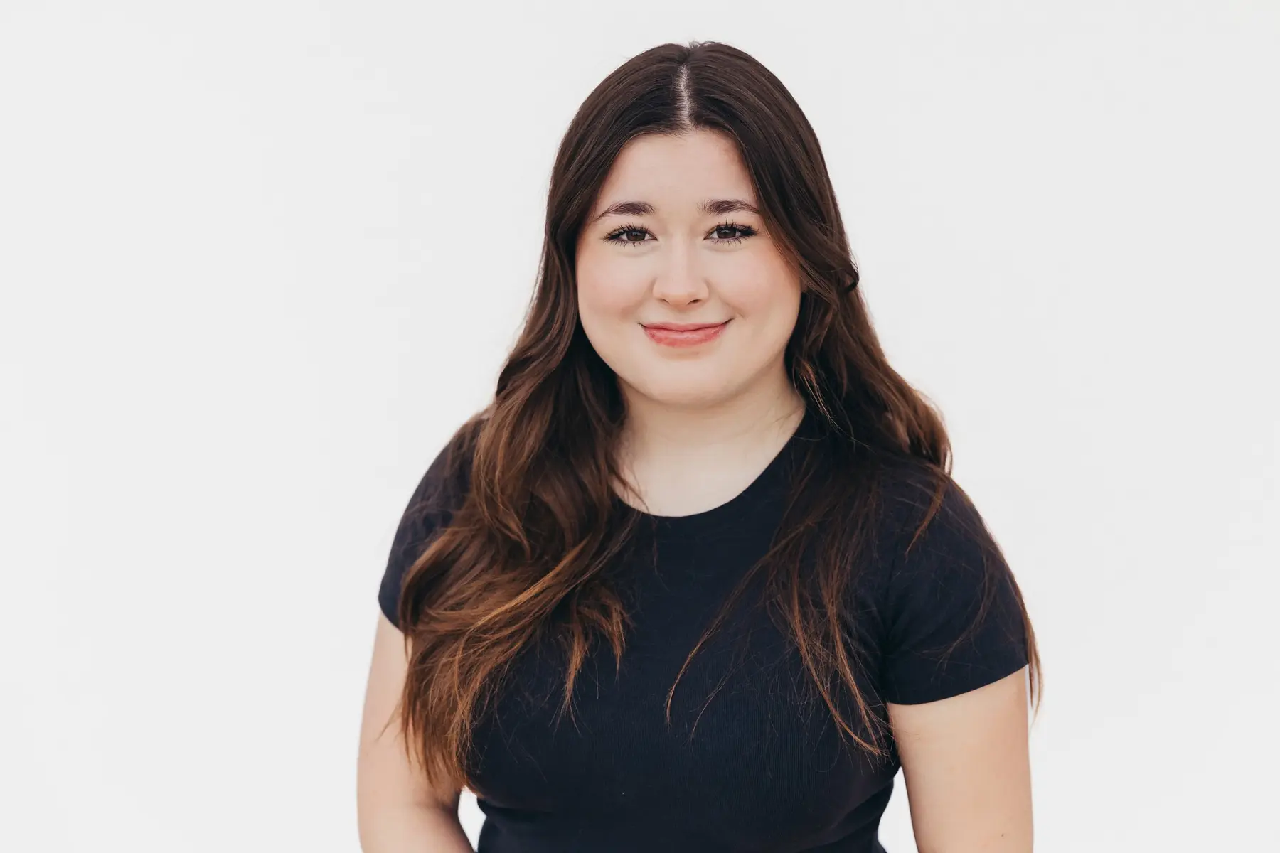A woman with long wavy brown hair wearing a black short-sleeved t-shirt smiles at the camera against a plain white background.