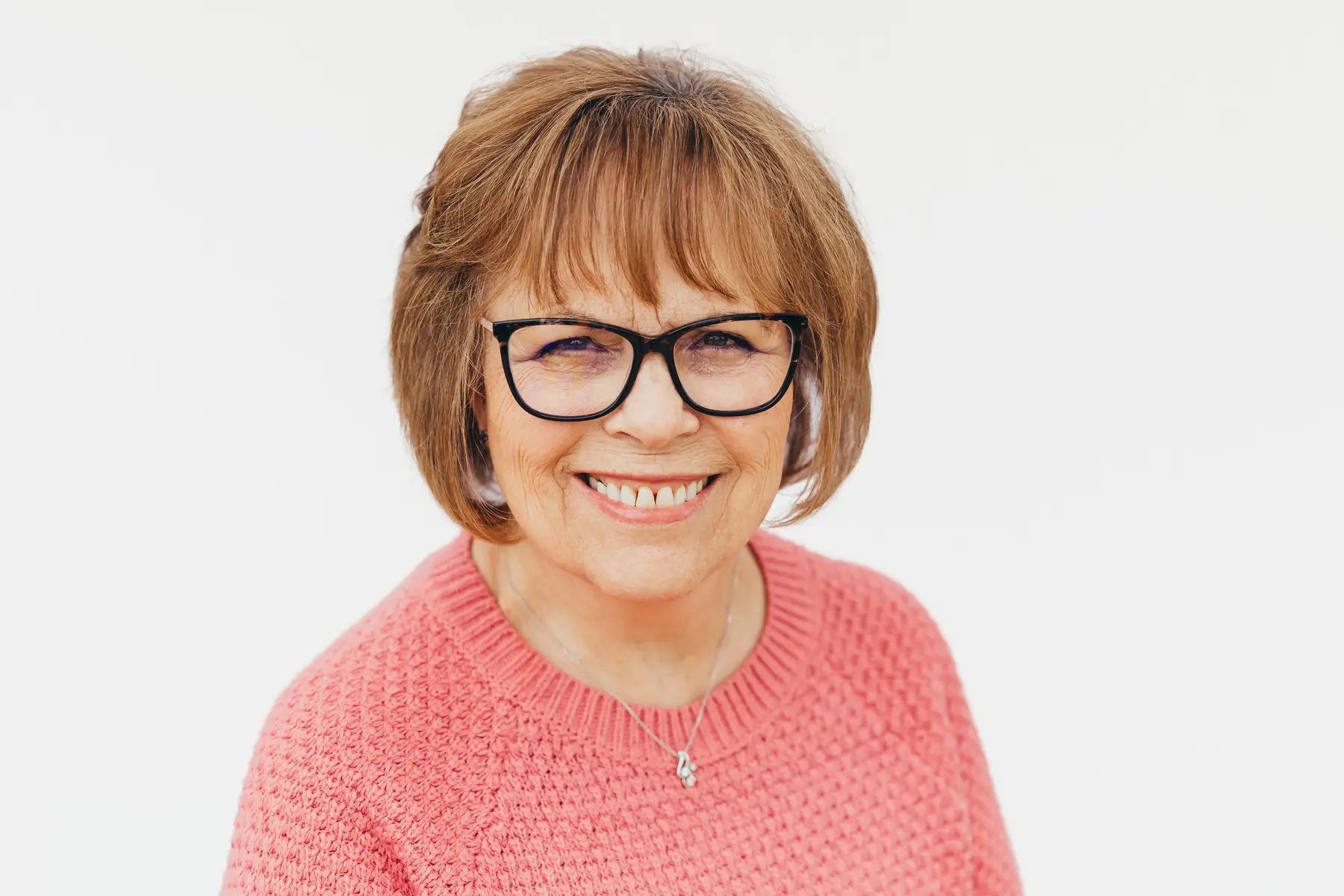 An older woman with short brown hair, black-rimmed glasses, and a pink knit sweater smiles at the camera against a plain white background.