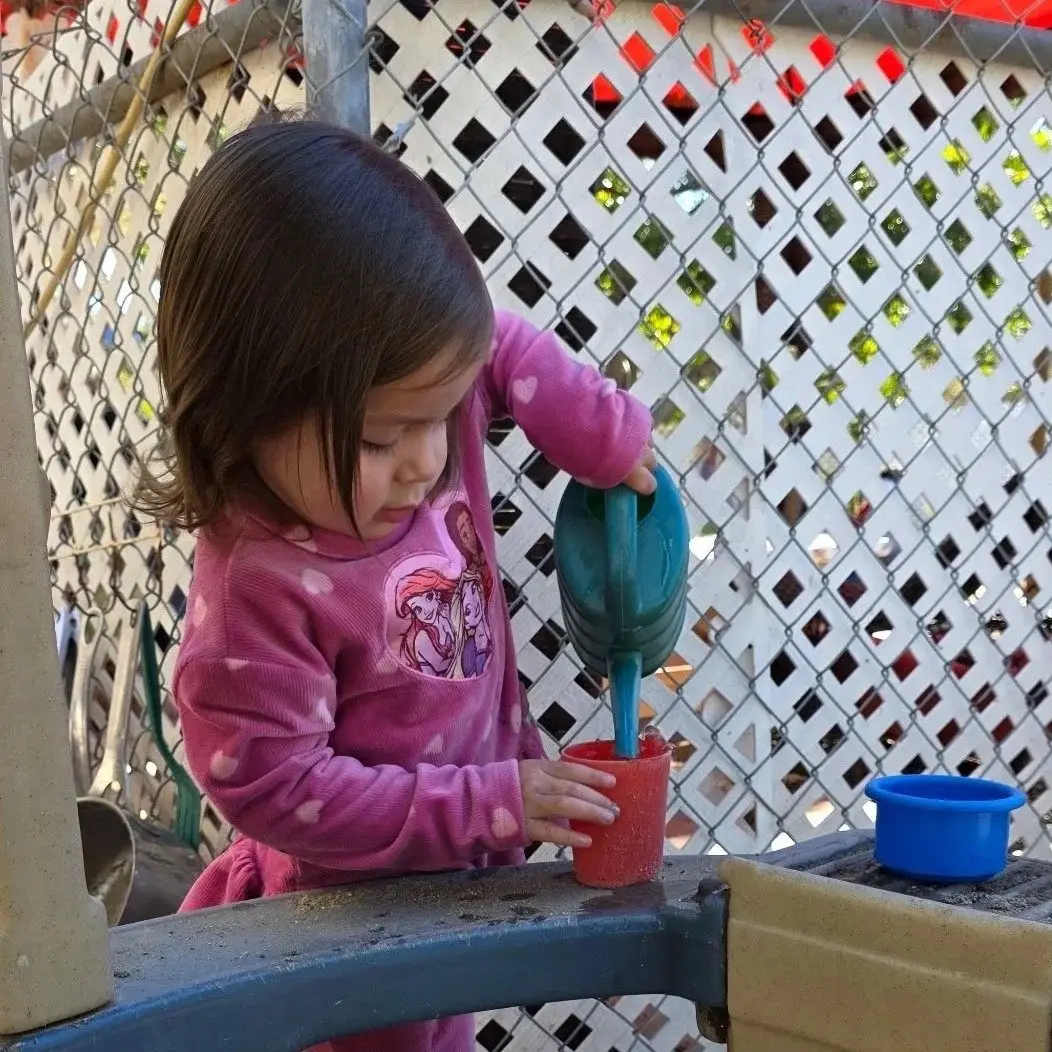 A young child in a pink heart-patterned shirt pours water from a teal watering can into a red cup on a play table.