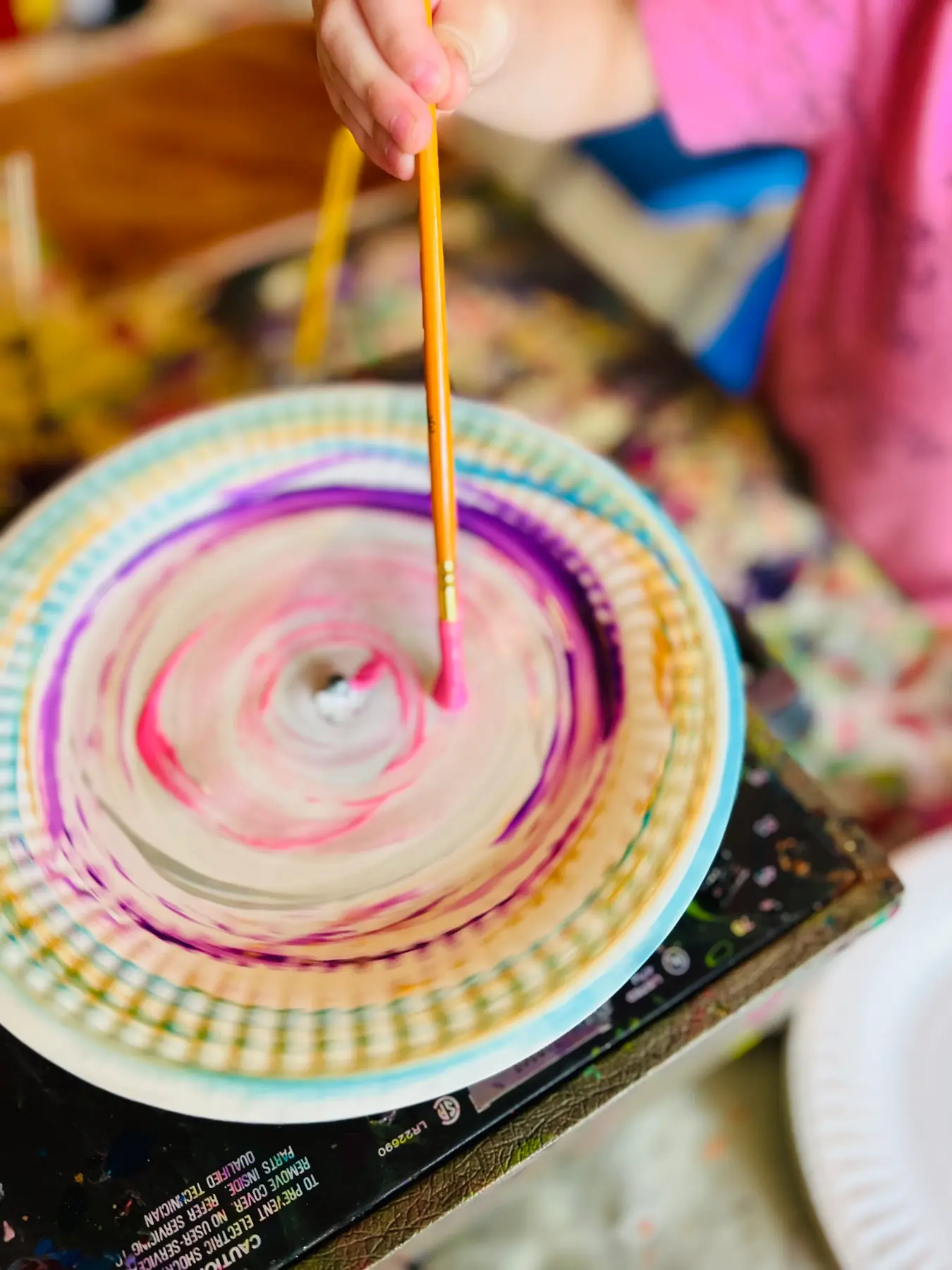 A child's hand holds a paintbrush and paints a swirling pink and purple design on a paper plate.