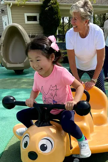 A young girl in a pink shirt sits on a yellow ride-on toy with a black steering wheel while an older woman in a white shirt stands behind her smiling.