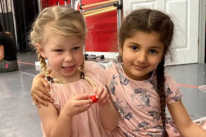 Two young girls sit close together indoors, one in a pink dress holding a small red object while the other in a floral dress rests an arm around her shoulder.
