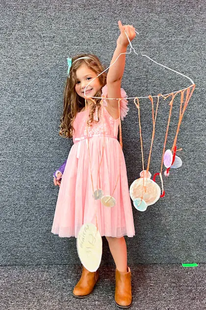 A young girl in a pink dress and brown boots stands holding up a wire hanger with hanging paper crafts against a gray textured wall.