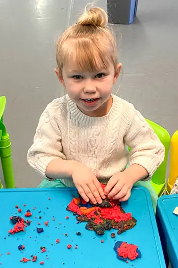 A girl in a cream sweater sits at a blue table and presses her hands into red and dark play dough.