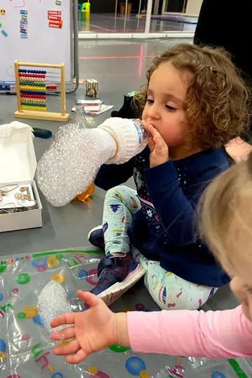 A toddler in a blue top and patterned leggings drinks from a bottle wrapped in bubble wrap while another child reaches toward a bubble-wrap-covered object on a table.