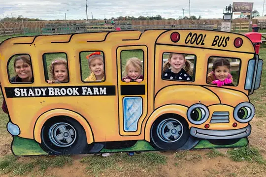 Five children and one adult pose inside a painted yellow school bus cutout at a farm.