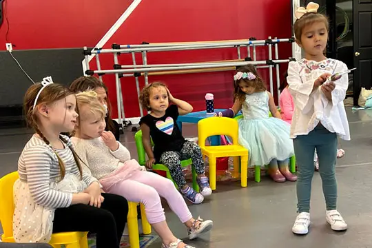 Several young girls sit on small yellow and green chairs while one girl stands holding a small object in front of them in a room with a red wall and ballet barre.