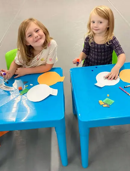 Two young girls sit at blue tables working on paper crafts with markers and foam shapes.